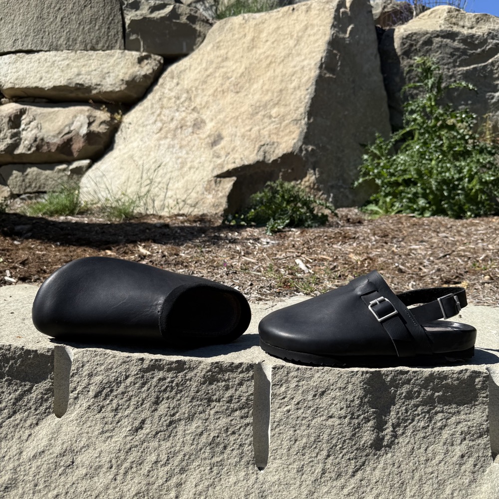 Handmade black leather sandals, one slip-on and one with a buckled slingback strap, on a stone wall