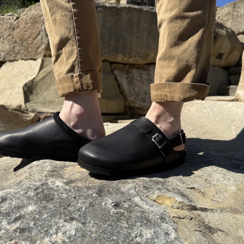 A pair of handmade black leather sandals worn with rolled khaki trousers on a rock
